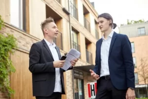 建物の前でスーツ姿の2人の男性が書類を手に立ち話をしている様子。