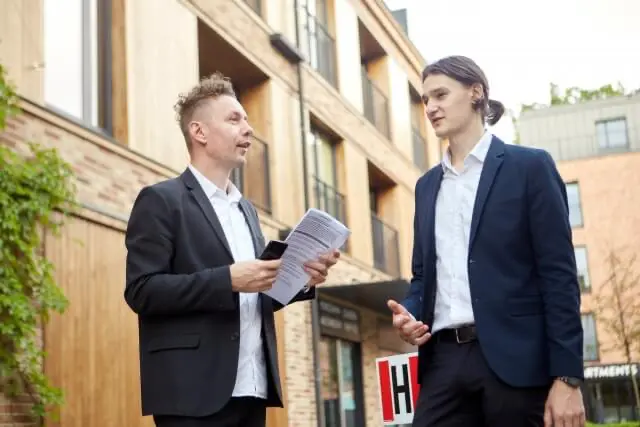 建物の前でスーツ姿の2人の男性が書類を手に立ち話をしている様子。