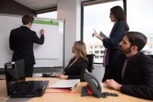 会議室でホワイトボードに書き込みながら説明する男性と、それを囲んで議論するビジネスパーソン達の様子