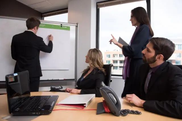 会議室でホワイトボードに書き込みながら説明する男性と、それを囲んで議論するビジネスパーソン達の様子