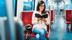 電車の通路側の赤い席に座って、黒い表紙カバーの本を読んでいる黒縁メガネをかけている女性の様子
