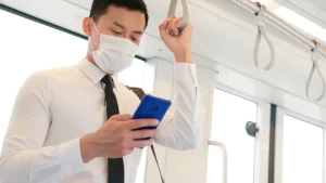 電車に吊り革につかまってスマートフォンを操作するマスクをしたネクタイ姿のビジネスパーソンの様子
