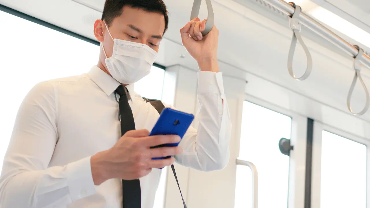 電車に吊り革につかまってスマートフォンを操作するマスクをしたネクタイ姿のビジネスパーソンの様子