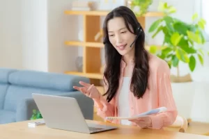 PCの前に座って、ヘッドフォンを装着して、左手に書類を持ち、オンラインで会話をする女性の様子