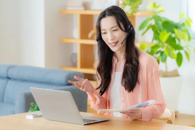 PCの前に座って、ヘッドフォンを装着して、左手に書類を持ち、オンラインで会話をする女性の様子