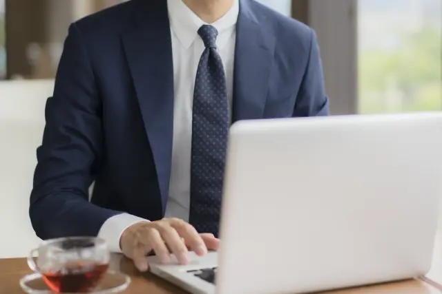 ノートPCに向かって仕事をしているネイビーのスーツ姿の男性の様子。ノートPCの横には紅茶が置かれている。
