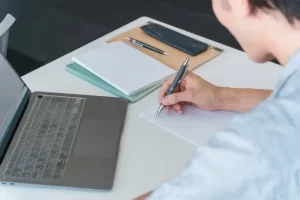 シルバーのボールペンでノートに書き込みながらパソコンで学習する男性のデスク作業風景