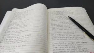 英文でぎっしり書かれたノートが開かれ、右ページには黒いペンが置かれている学習中の様子